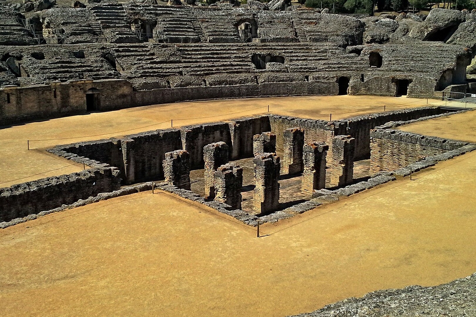 The image shows the well-preserved ruins of the Roman Amphitheatre of Itálica, located in modern-day Santiponce, near Seville, Spain.