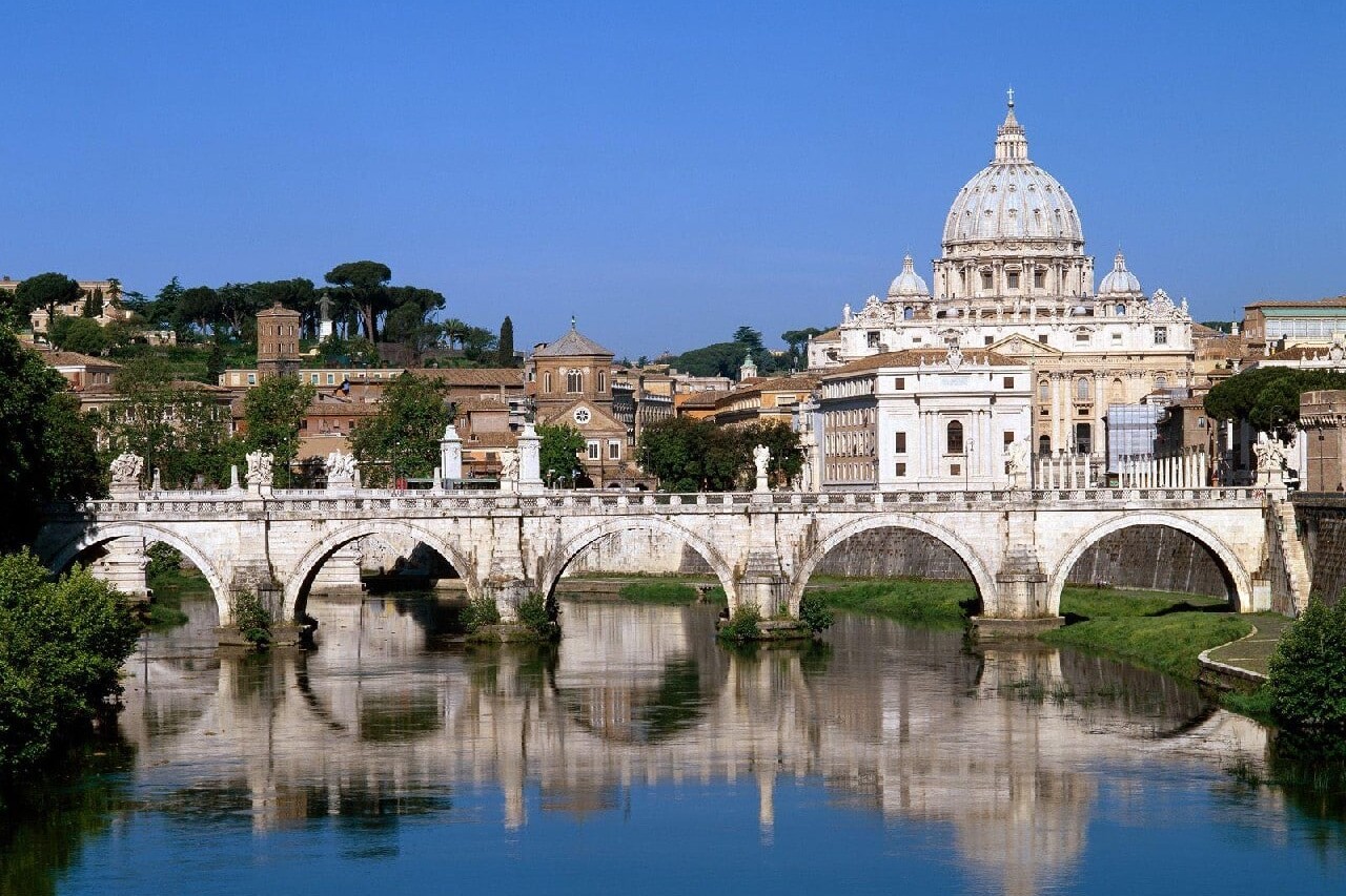The image displays the Ponte Sant'Angelo bridge over the Tiber River with St. Peter's Basilica in the background, located in Vatican City.