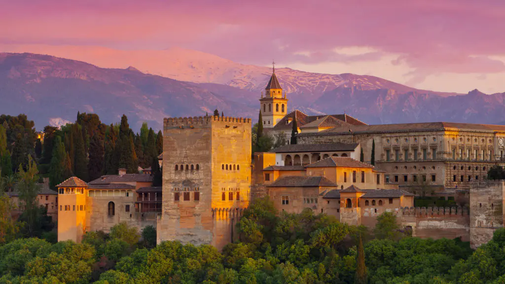 The Alhambra palace glows at dusk above Granada, its Nasrid towers and defensive walls lit against the snowcapped Sierra Nevada mountains behind