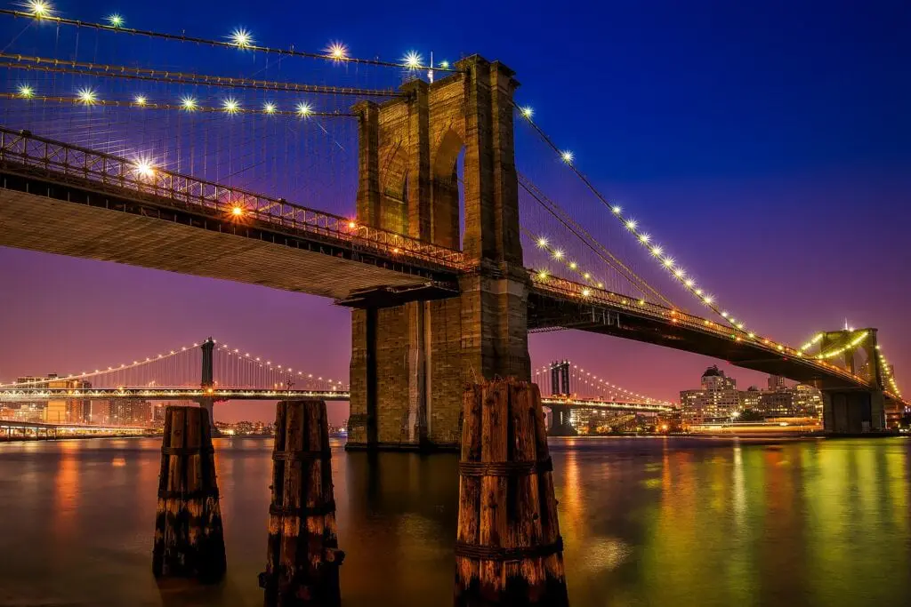 The Brooklyn Bridge spans the East River at dusk, its stone towers and steel cables framing the glittering Manhattan skyline beyond