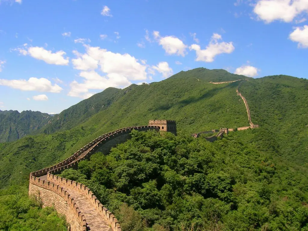 The Great Wall of China winds through vibrant green summer mountains, disappearing into the hills toward the horizon