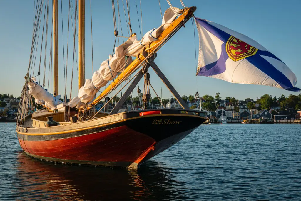 A traditional wooden schooner flies the Nova Scotia flag in Lunenburg Harbor, with the UNESCO-listed town's colorful waterfront visible across the water